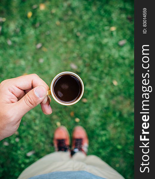 Closeup of a hand holding a cup of black coffee and selective focus blurring blue jumper, beige trousers and pink trainers or shoes. Closeup of a hand holding a cup of black coffee and selective focus blurring blue jumper, beige trousers and pink trainers or shoes.