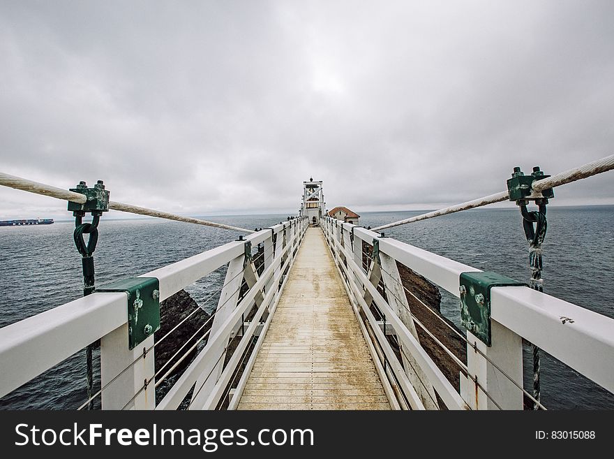 White suspension bridge with wooden planking floor showing supporting cables and safety wires, background of gray cloudy sky. White suspension bridge with wooden planking floor showing supporting cables and safety wires, background of gray cloudy sky.