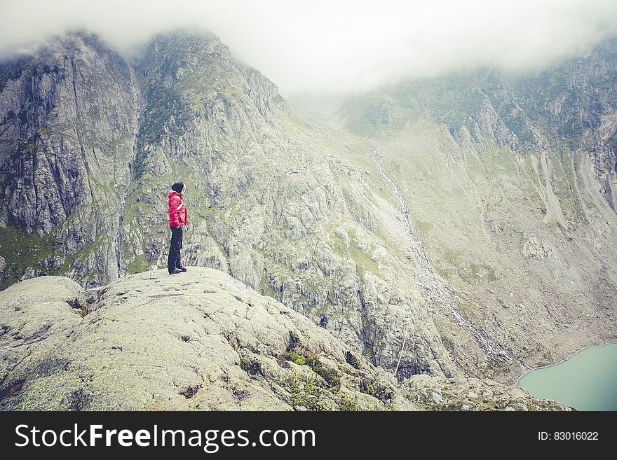 Character in red jacket and dark trousers standing on mountain top looking down upon blue Lake. Tops of nearby mountain range shrouded in fog and mist. Concept of getting to the top or great achievement. Character in red jacket and dark trousers standing on mountain top looking down upon blue Lake. Tops of nearby mountain range shrouded in fog and mist. Concept of getting to the top or great achievement.