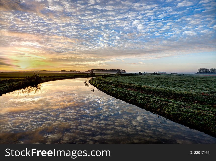 Clouds reflecting in river through green fields at sunset.