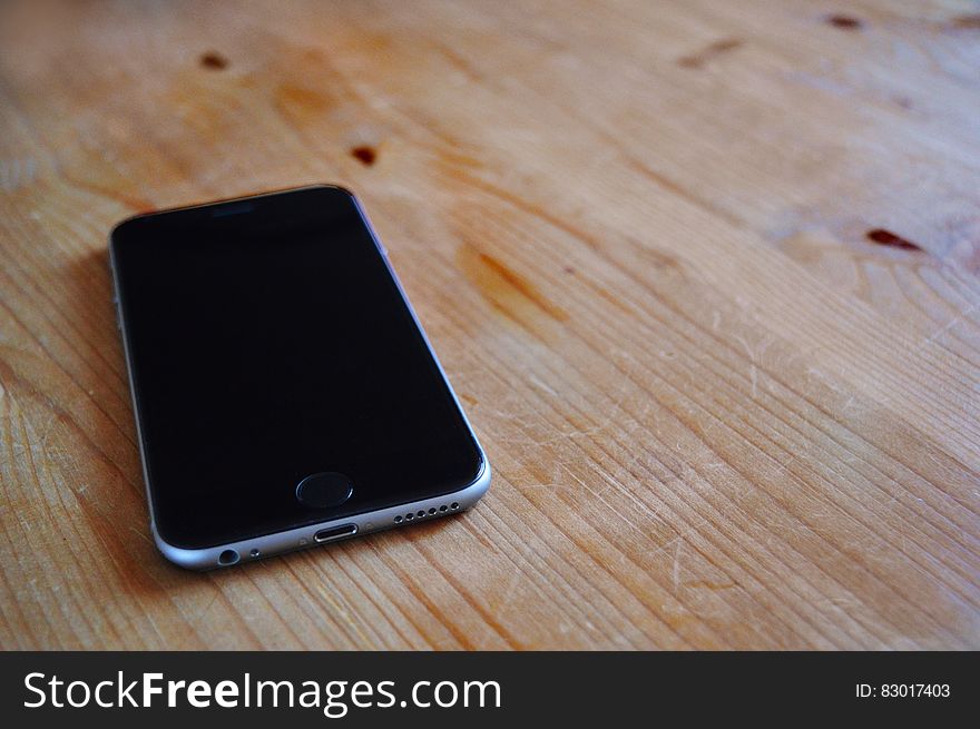 Apple iPhone with blank black screen and silver (or chrome) casing showing charger socket, speaker and headphone socket, lying upon grainy wooden table.