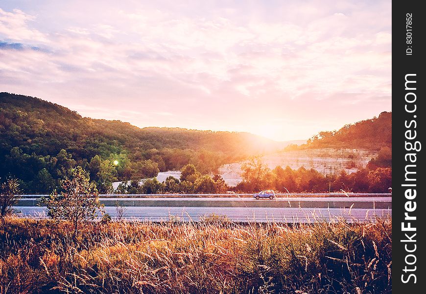 Sunrise over interstate highway with a single small car on the road and golden countryside with forests to left and right.