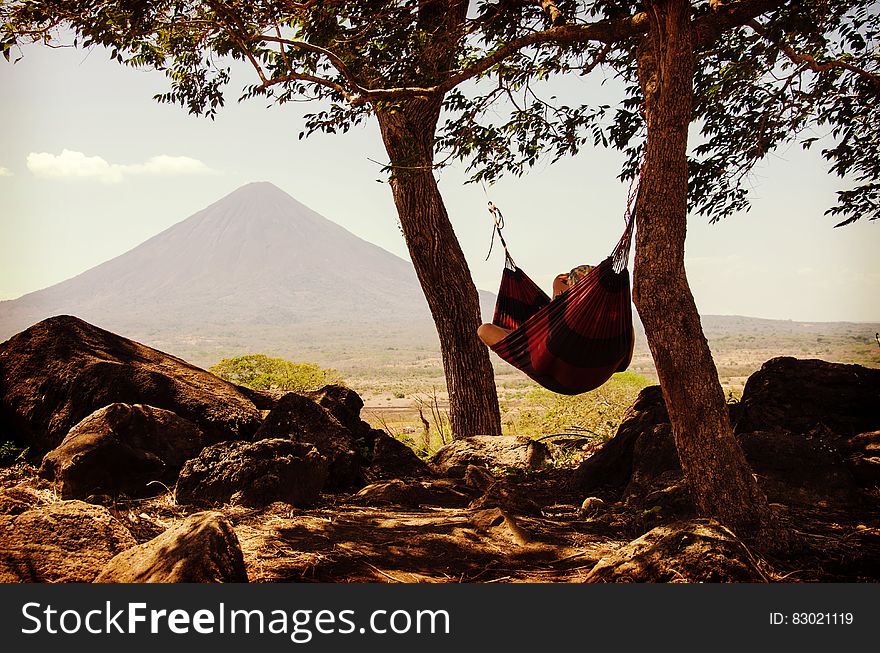 Person Lying On Black And Red Hammock Beside Mountain Under White Cloudy Sky During Daytime