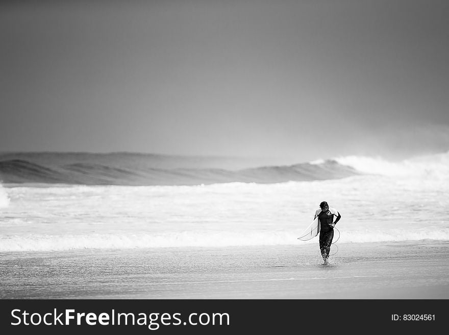 Surfer in wet suit leaving the sea and walking up the beach after surfing on big breaking waves. Surfer in wet suit leaving the sea and walking up the beach after surfing on big breaking waves.