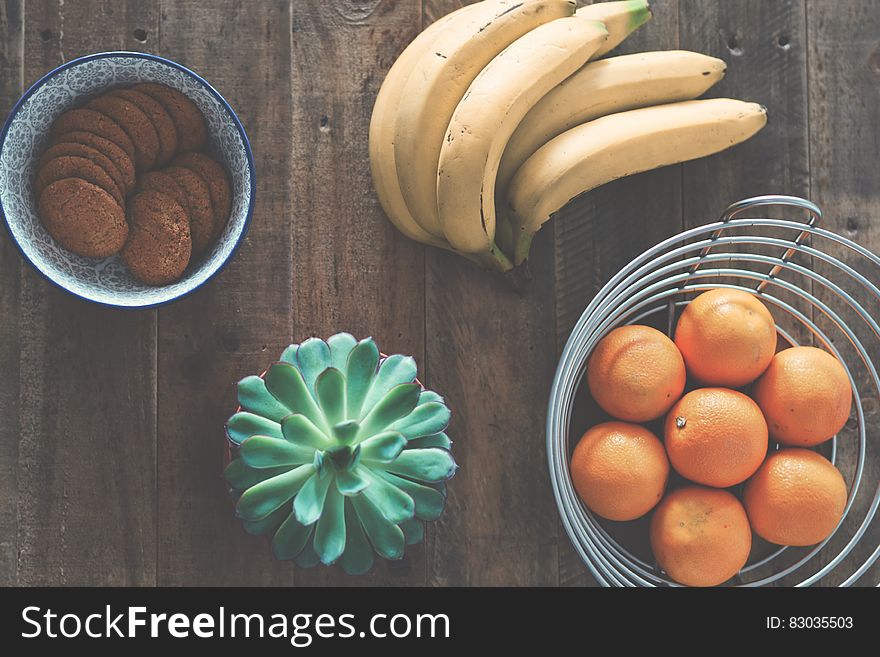 Fresh bananas and wire basket of oranges on wooden tabletop with bowl of cookies. Fresh bananas and wire basket of oranges on wooden tabletop with bowl of cookies.