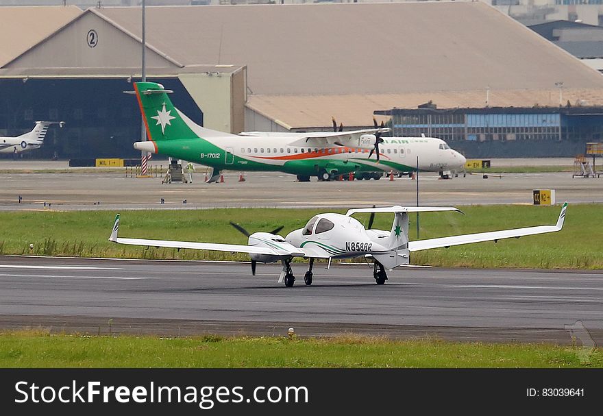 Airport with large green, white and red propeller driven passenger aircraft and smaller white private plane on the runway, hangars in the background. Airport with large green, white and red propeller driven passenger aircraft and smaller white private plane on the runway, hangars in the background.