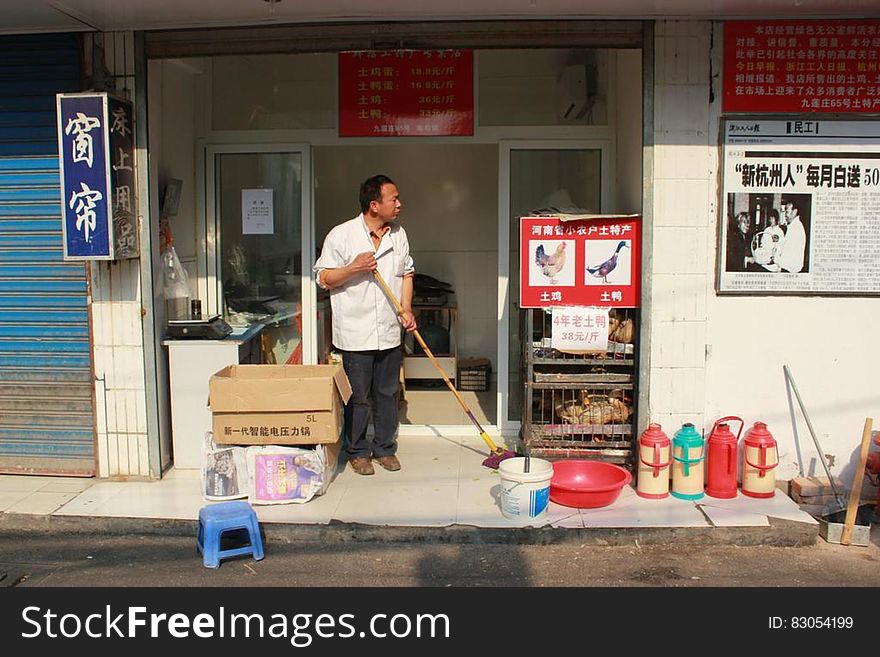 Asian shopkeeper sweeping storefront on sunny day.