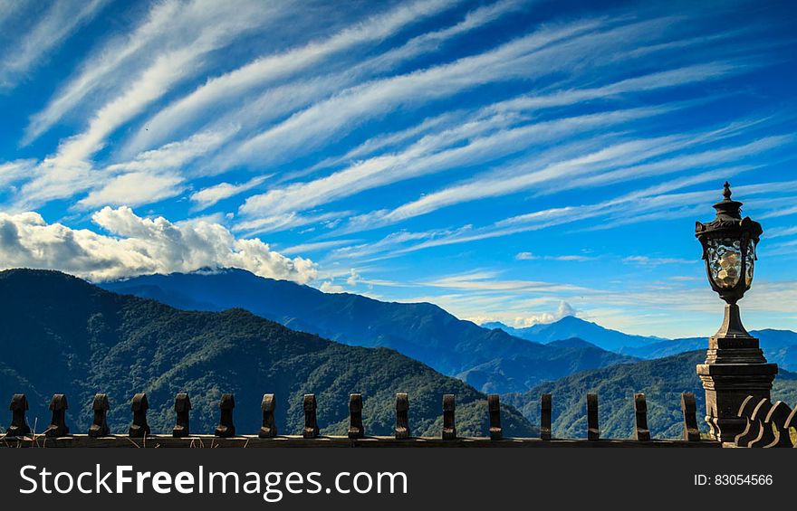 Dramatic white clouds in blue skies over mountain range above metal fencing and lamppost. Dramatic white clouds in blue skies over mountain range above metal fencing and lamppost.