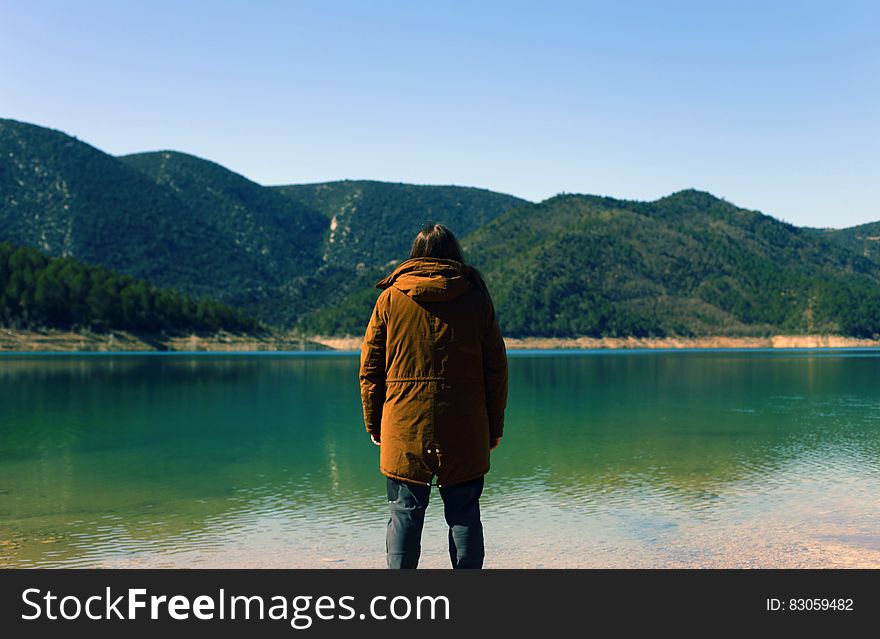 Man wearing brown jacket with hood, dark trousers and boots looking across tranquil lake towards distant mountains. Man wearing brown jacket with hood, dark trousers and boots looking across tranquil lake towards distant mountains.