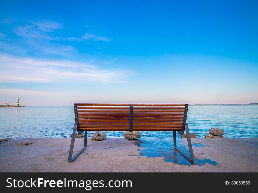 Empty bench on waterfront against blue skies on sunny day.