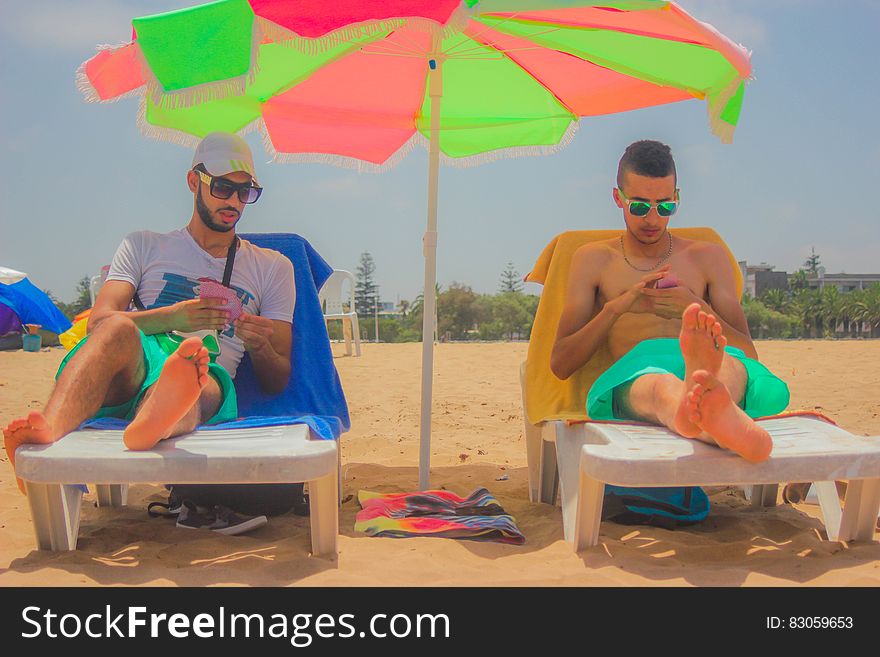 2 Man Sitting on White Beach Lounger With Green and Pink Patio Umbrella in Between