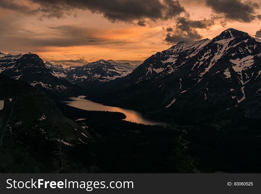 Sunset over mountain lake with snowy peaks and cloudy skies.