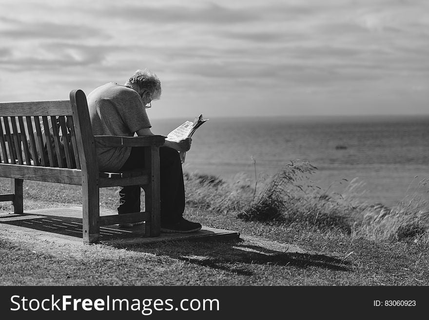 Grayscale Photo of Man Sitting on Brown Wooden Bench Reading News Paper during Day Time