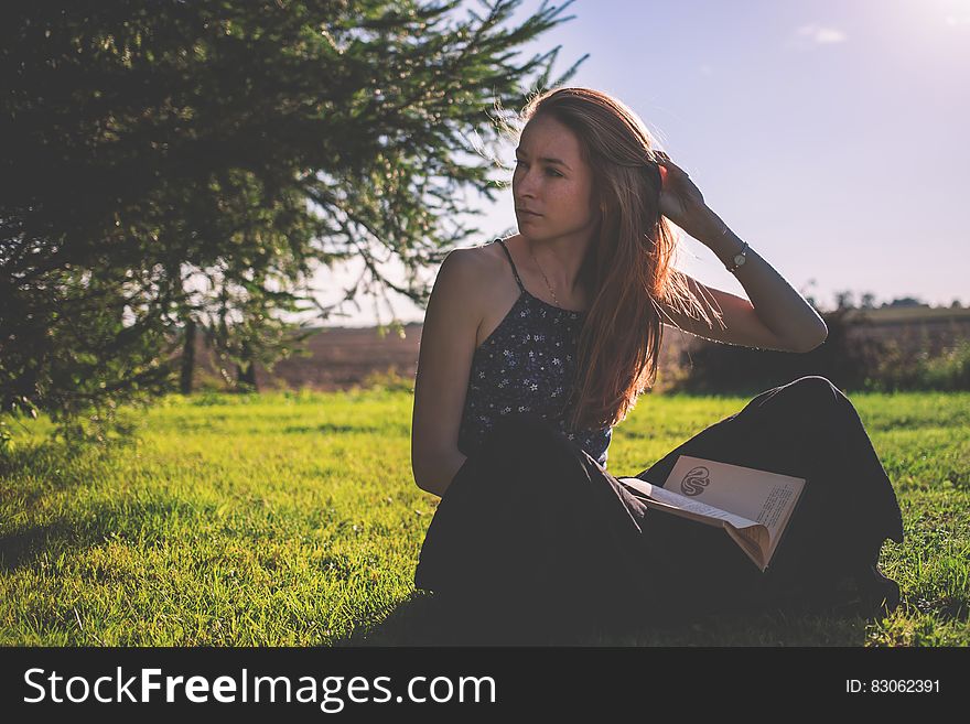 Woman in Black Tank Top and Holding Brown Book Sitting on Grass Under Clear Sunny Sky