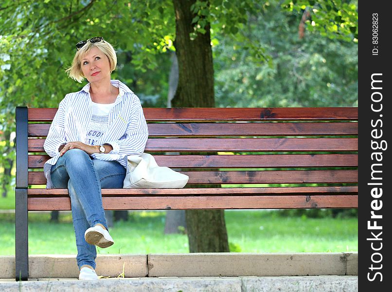 Thinking Woman in White Jacket and White Scoop Neck Shirt Blue Denim Jeans Sitting on Brown Wooden Bench Beside Green Trees during