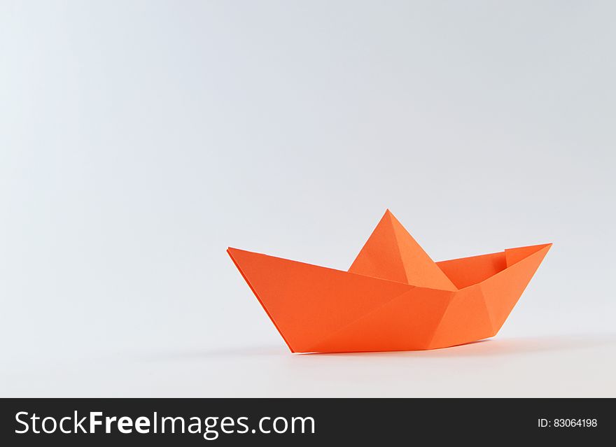 An orange colored stylized boat made by origami, that is folding plain orange paper, white background. An orange colored stylized boat made by origami, that is folding plain orange paper, white background.