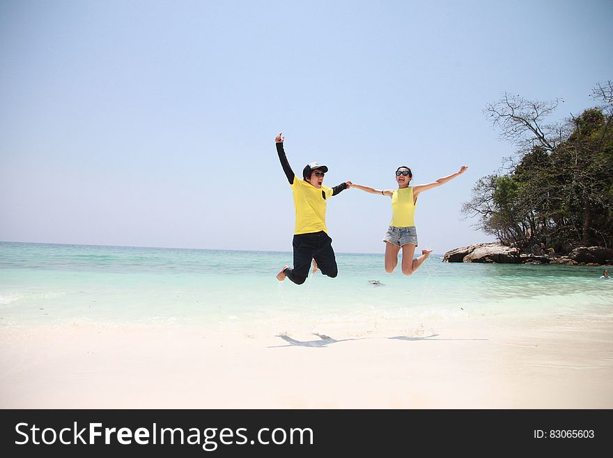 Woman in Blue Denim Mini Short Smiling While Holding Another Person in a Jump Shot Photo at Seashore during Daytime