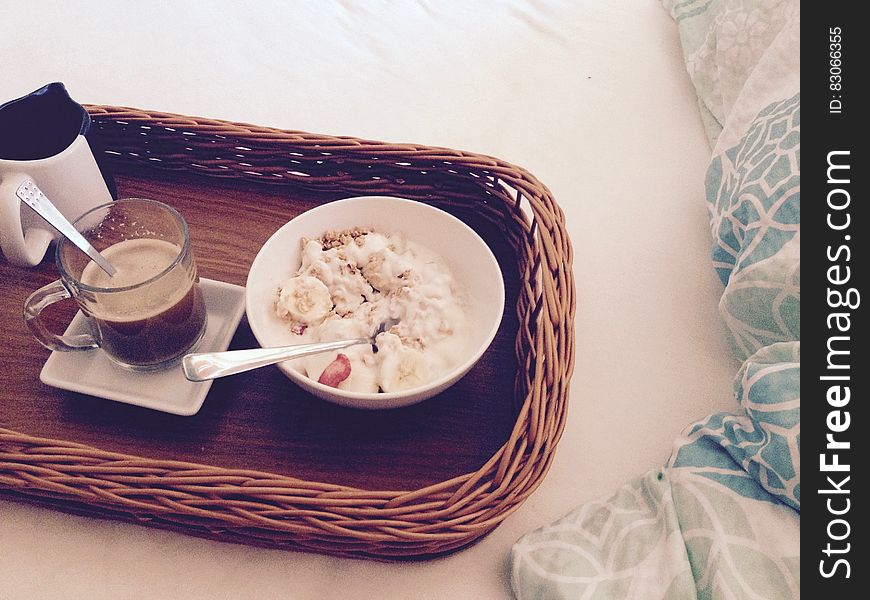 White Round Bowl With Rice And Sliced Bananas Near Clear Glass Mug On Square Ceramic Plate And White And Black Ceramic Mug On Brow