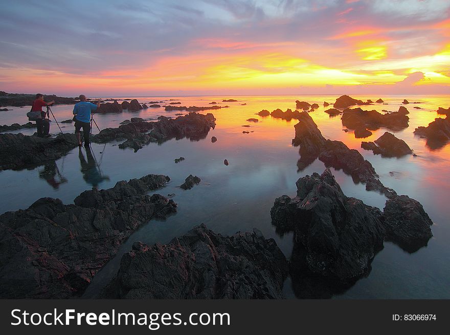 2 People Standing On Rock Formation Reflected On Water During Sunset