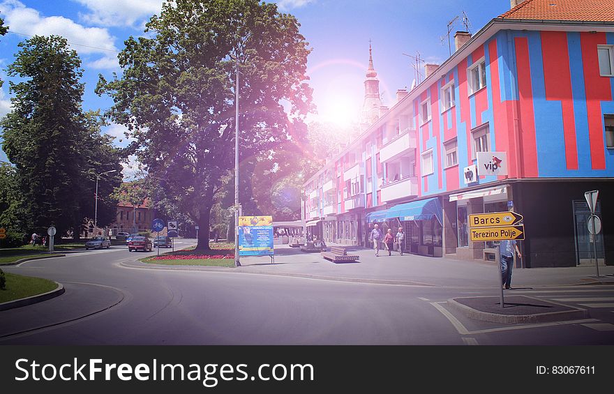 Unusual urban architecture with red and blue paneling in apartments above shops, orange tiled roofs and tree lined road.