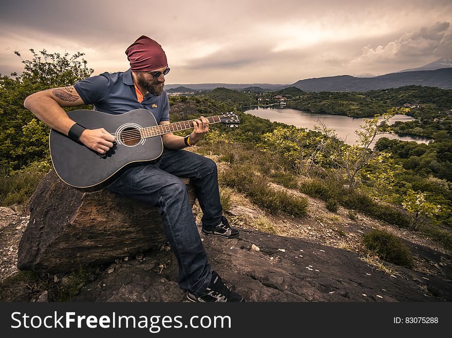 Portrait of man in bandanna playing guitar sitting on hilltop. Portrait of man in bandanna playing guitar sitting on hilltop.
