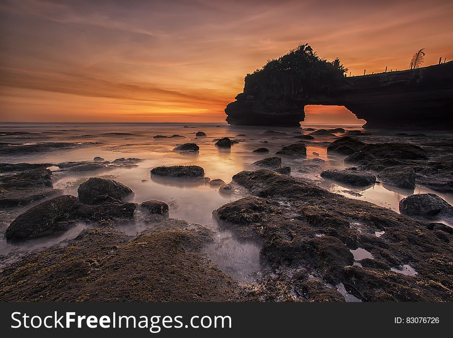 Orange And Yellow Sunset Skies Over Grey Seashore Rocks And Calmed Ocean