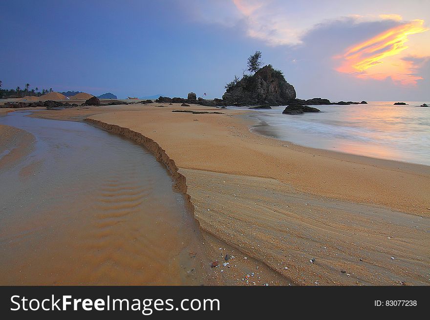 Beach at sunset with receding tide and rocky protrusion with tree growing on it. Beach at sunset with receding tide and rocky protrusion with tree growing on it.