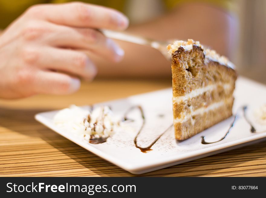 A close up of a piece of cake on a white plate and a hand holding a fork reaching for it.