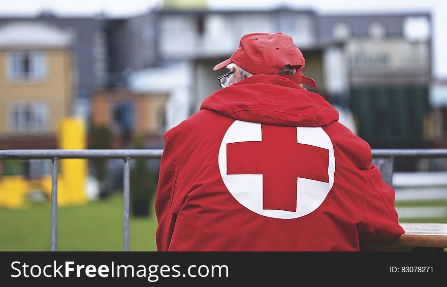 An elderly man in a red cross jacket next to a metal fence. An elderly man in a red cross jacket next to a metal fence.