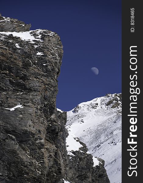 Mountain rocks partially covered in snow with blue sky and moon. Mountain rocks partially covered in snow with blue sky and moon