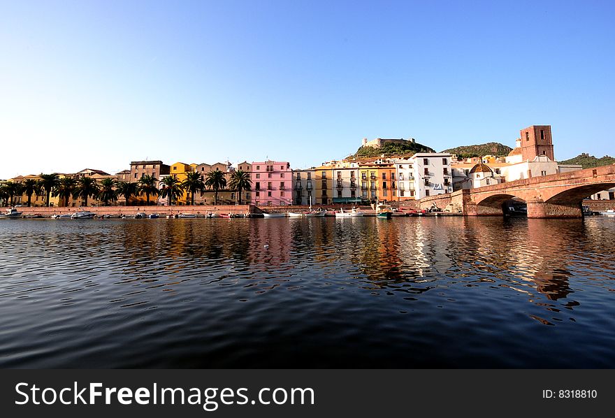 Colourful Mediterranean town of Bosa Sardinia with buildings and homes running along the river banks with the clear still water in front reflecting image back and old stone bridge to the side. Colourful Mediterranean town of Bosa Sardinia with buildings and homes running along the river banks with the clear still water in front reflecting image back and old stone bridge to the side.