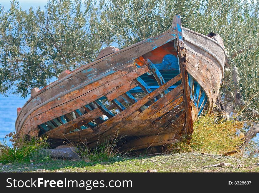 A skeletal blue and orange boat abandoned in an olive orchard on a mountain with blue sea behind. A skeletal blue and orange boat abandoned in an olive orchard on a mountain with blue sea behind