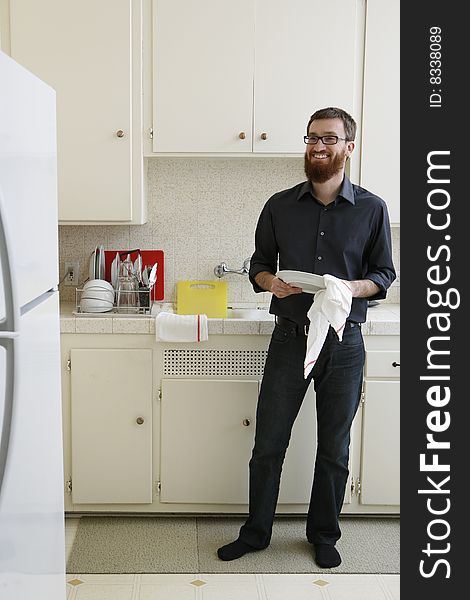 Man with beard smiles while helping in the kitchen.  He is wearing a dark shirt and denim is standing by sink. Man with beard smiles while helping in the kitchen.  He is wearing a dark shirt and denim is standing by sink.