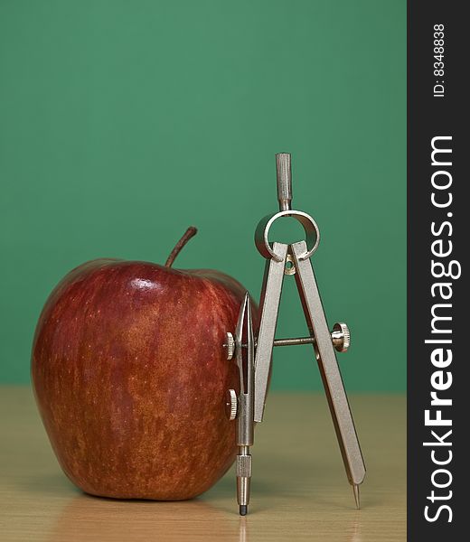 A red apple and a drawing compass over a classroom desk. Blank chalkboard on the background. A red apple and a drawing compass over a classroom desk. Blank chalkboard on the background.