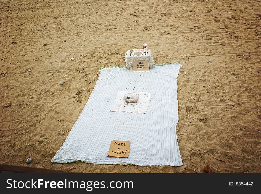 A panhandlers blanket, on the beach, with a dinner palcesetting, set up to accept donations. A panhandlers blanket, on the beach, with a dinner palcesetting, set up to accept donations.