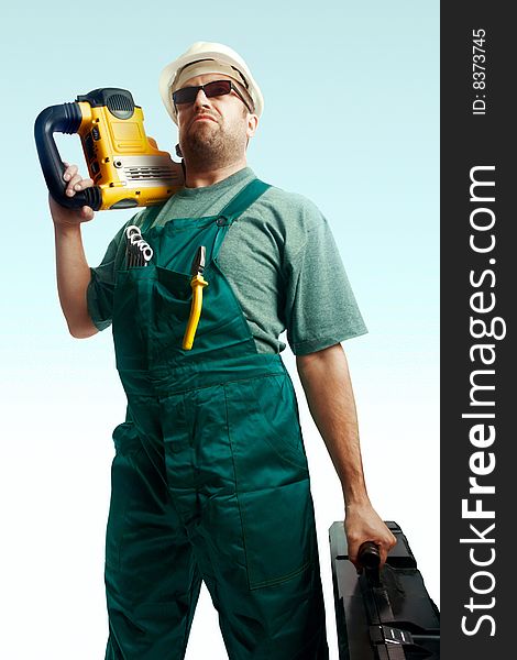 Serious skeptic workman in helmet, black glasses and overalls hold perforator on the shoulder and big black suitcase over white background. Serious skeptic workman in helmet, black glasses and overalls hold perforator on the shoulder and big black suitcase over white background