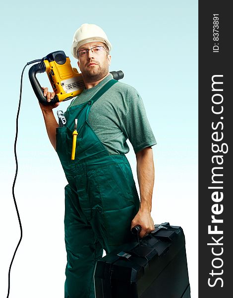 Serious workman in helmet, glasses and overalls hold perforator on the shoulder and big black suitcase in hand over white background. Serious workman in helmet, glasses and overalls hold perforator on the shoulder and big black suitcase in hand over white background