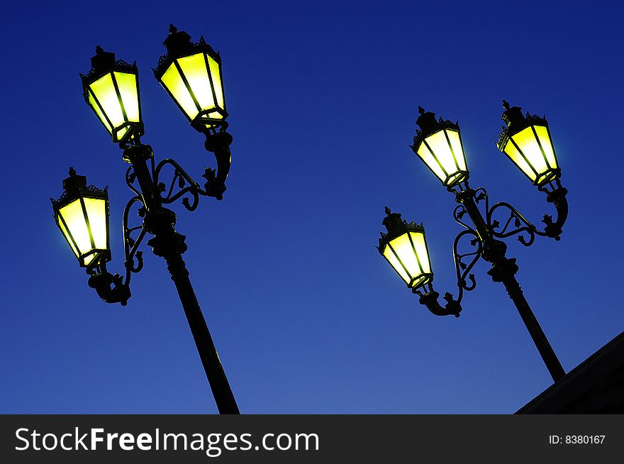 Two sloped, street lights in night dark blue sky. Yellow luminescence. Two sloped, street lights in night dark blue sky. Yellow luminescence.