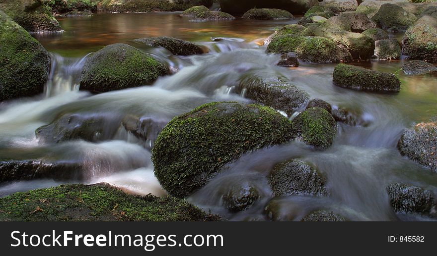 A wide shot of a stream with the motion of the water blurred around moss covered rocks and stones. A wide shot of a stream with the motion of the water blurred around moss covered rocks and stones