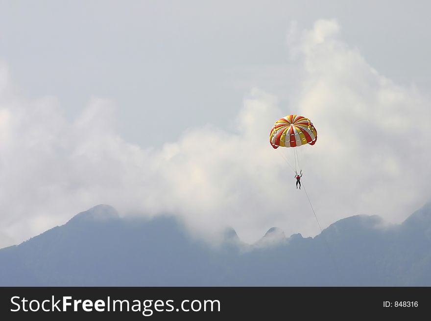 A male vacationer parasailing high in the sky above the mountains of Langkawi Island, Malaysia. The speedboat pulling him is omitted. A male vacationer parasailing high in the sky above the mountains of Langkawi Island, Malaysia. The speedboat pulling him is omitted.