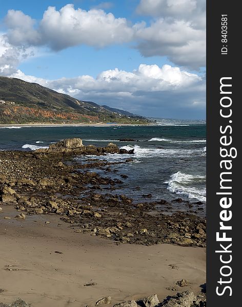The rocky shore and golden sands of Leo Carillo Beach in Southern California, with mountains in the background and white clouds in the sky. The rocky shore and golden sands of Leo Carillo Beach in Southern California, with mountains in the background and white clouds in the sky