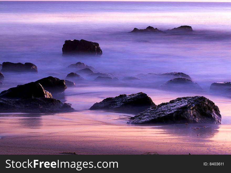 Rocks off the coast of Southern California sit in the mists of the rolling tides at sunset. The beautiful colors of the dusk sky are reflected in the turbulent waters. Rocks off the coast of Southern California sit in the mists of the rolling tides at sunset. The beautiful colors of the dusk sky are reflected in the turbulent waters
