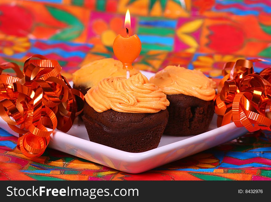 Chocolate cupcakes with orange frosting on a brightly colored background with a lit candle and ribbons. Chocolate cupcakes with orange frosting on a brightly colored background with a lit candle and ribbons