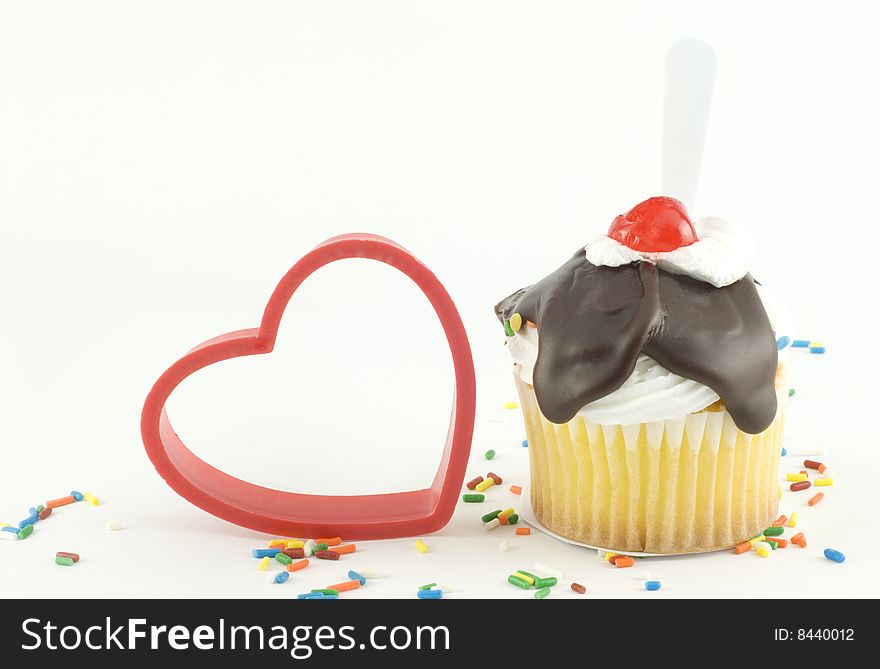 A cute sundae cupcake with yellow cake and chocolate fudge topping with cherry and sprinkles with red plastic heart on white background with copy space. A cute sundae cupcake with yellow cake and chocolate fudge topping with cherry and sprinkles with red plastic heart on white background with copy space