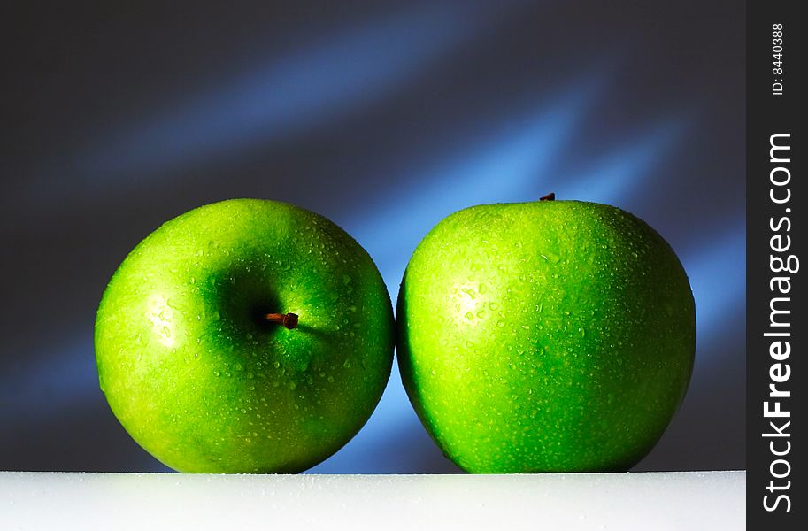 Two green apples on a white surface on a dark blue background. Are covered by water drops