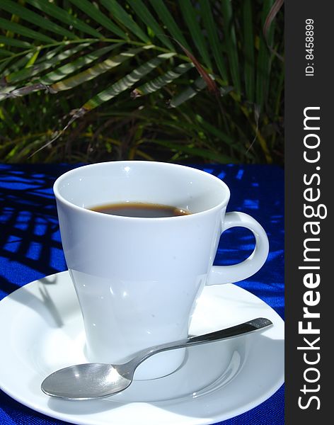 Black coffee in white cup with plate and spoon on blue and palms in the background