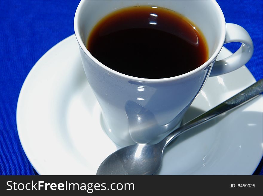 Black coffee in white cup with plate and spoon on blue background