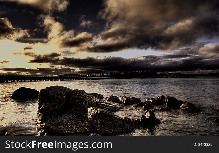 A brooding sunset over the sea with rocks in the foreground and causeway in the background.