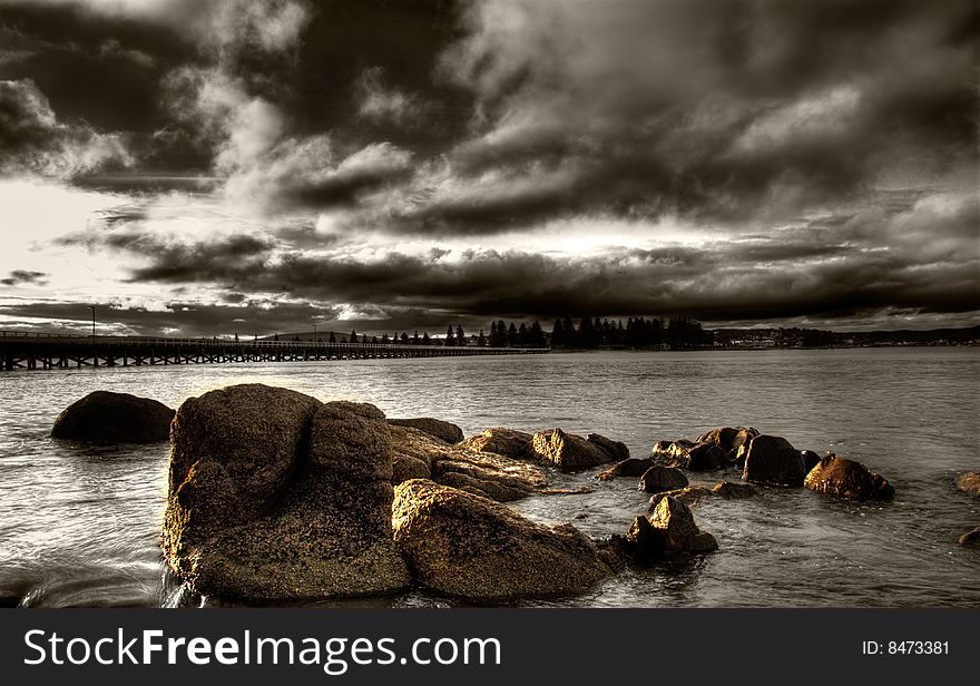 A brooding sunset over the sea with rocks in the foreground and causeway in the background.