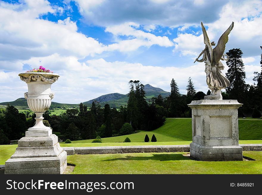 Formal garden with statues, mountains in background, blue sky and clouds